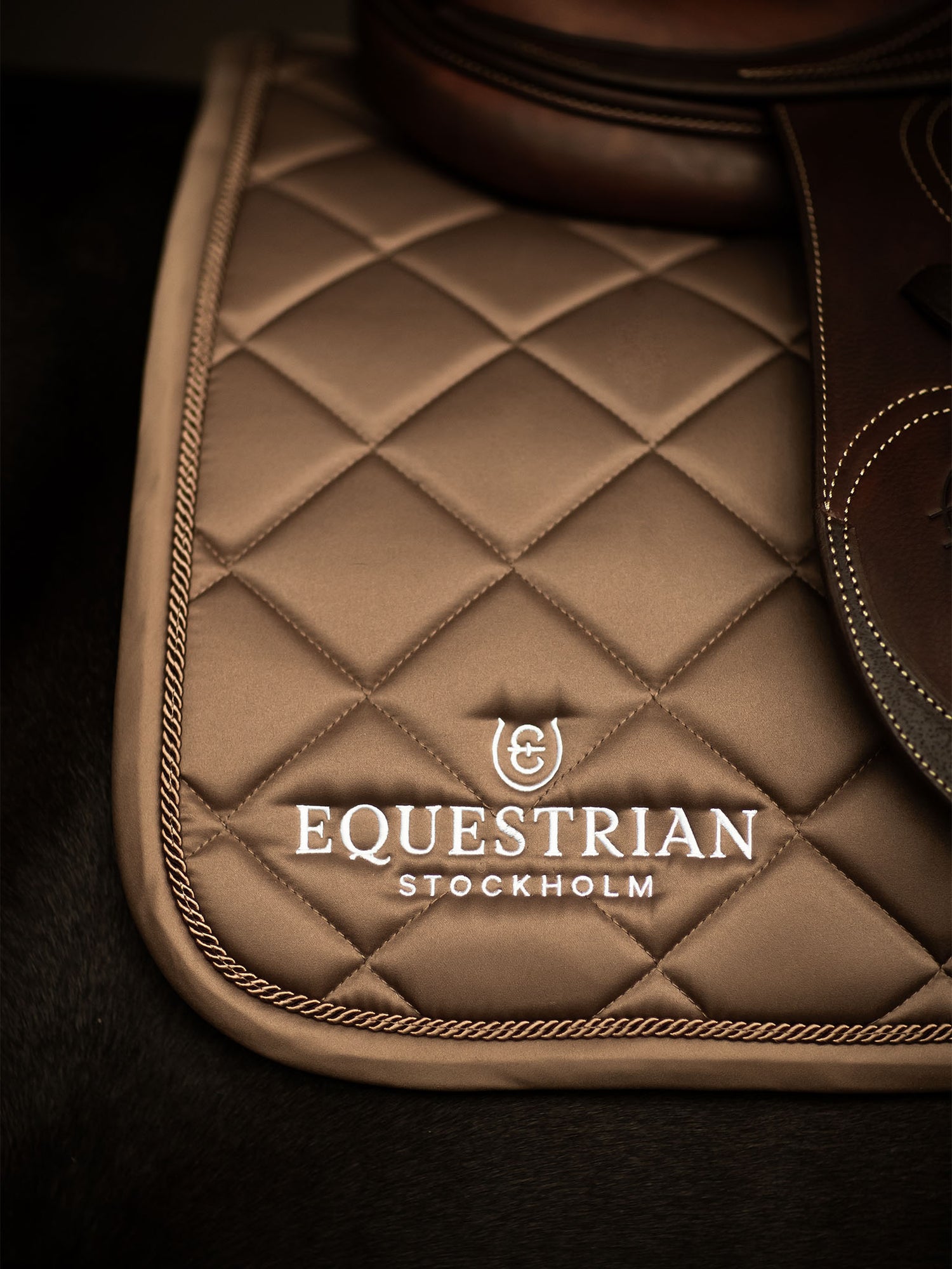 Studioaufnahme der Equestrian Stockholm Springschabracke Line Champagne mit Rautensteppung, weißem Logo und braunem Sattel.