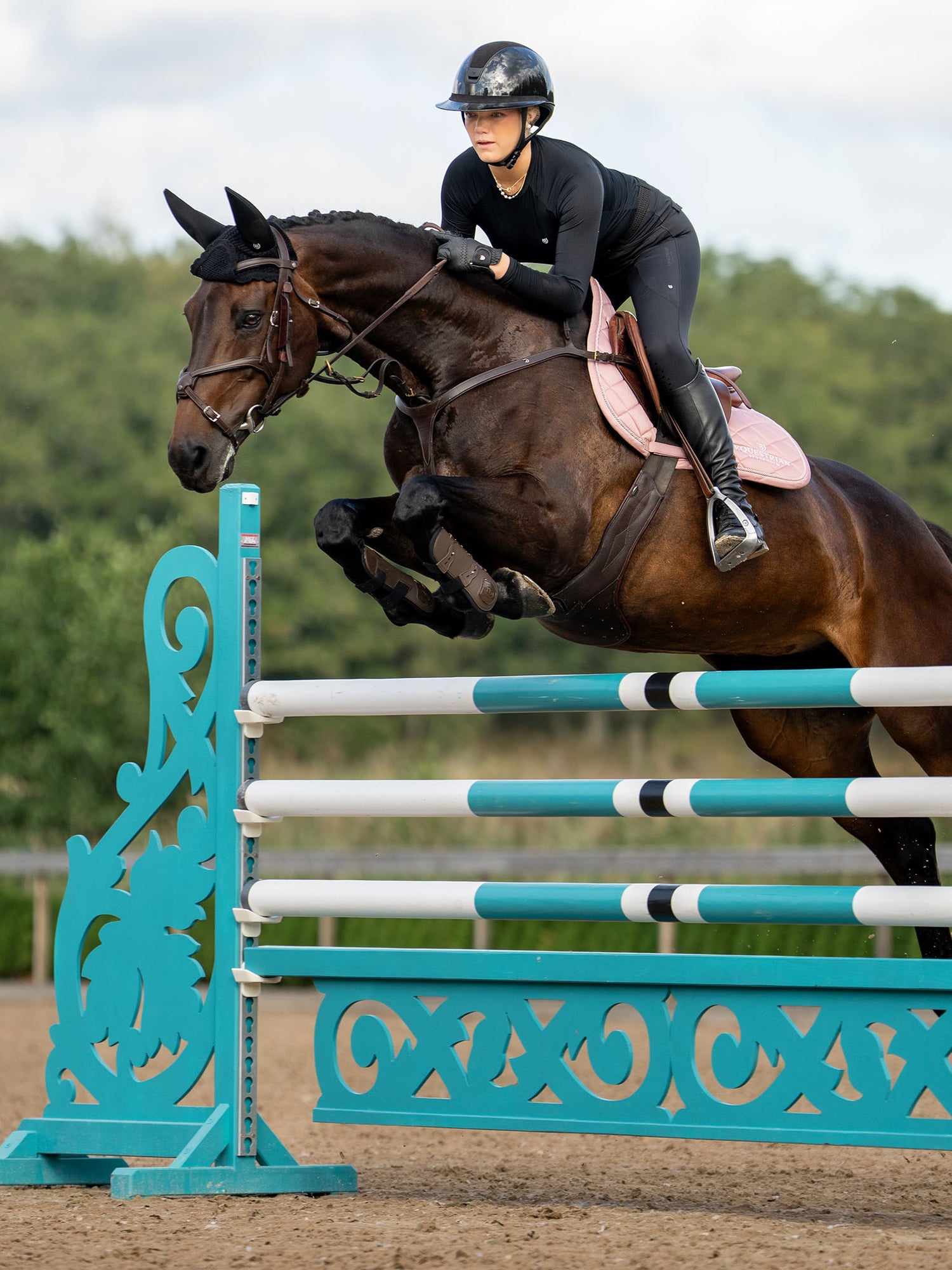A rider jumps a horse wearing the Equestrian Stockholm Jump Saddle Pad Line Pink over a teal and white hurdle.