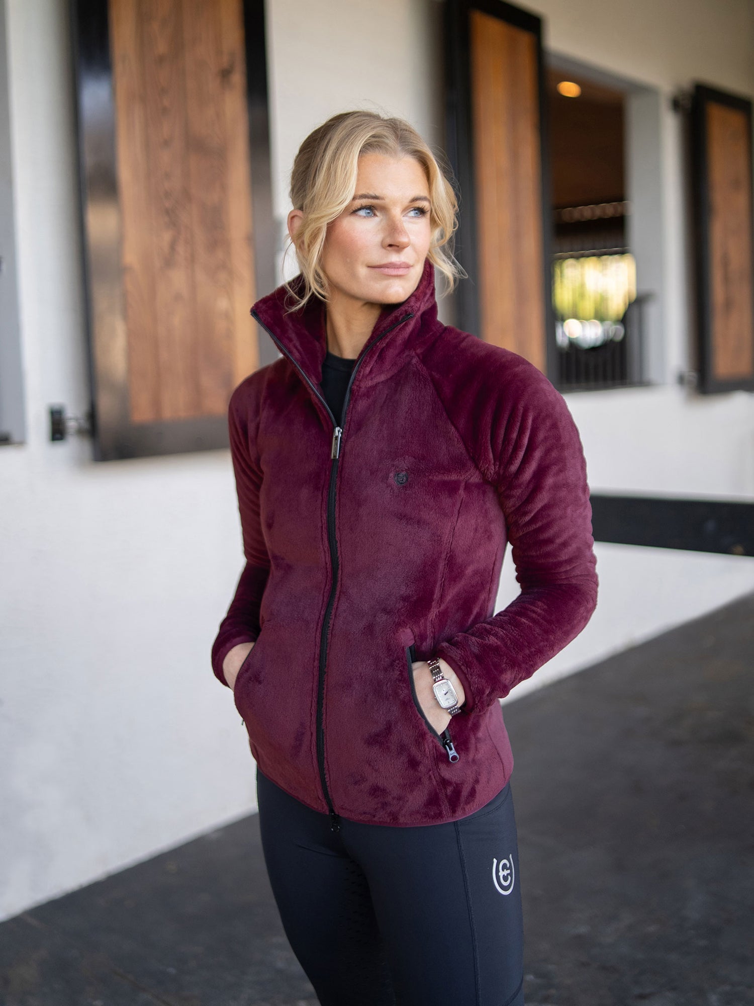 A model wearing a Merlot Equestrian Stockholm Velvet Fleece Jacket and black riding tights at a stable.