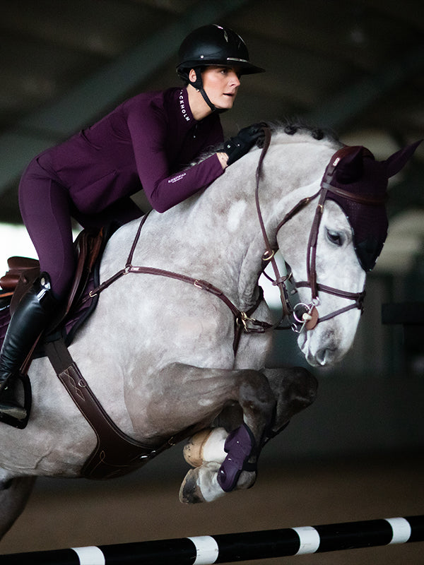 Reiter beim Sprung auf einem Schimmel in Equestrian Stockholm Black Raven Reitleggings, Top, Fliegenhaube und Gamaschen.