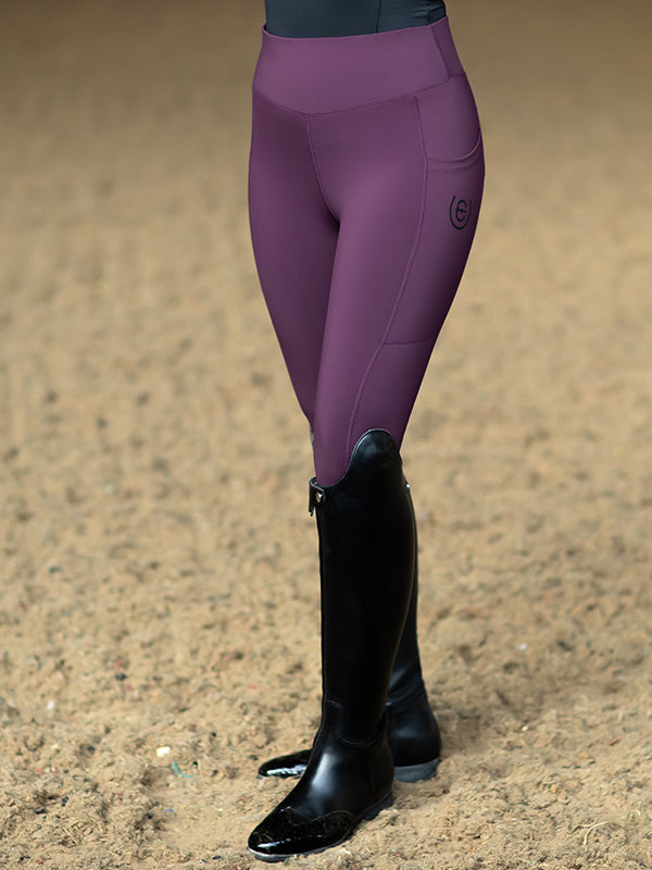 Studioaufnahme der Equestrian Stockholm Reitleggings Movement Black Raven in Dunkelviolett mit schwarzen Stiefeln auf Sand.