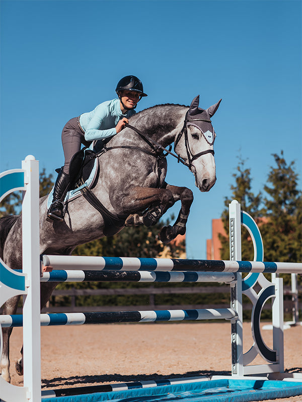 Sottosella da salto e cuffia per orecchie Ice Blue Equestrian Stockholm su un cavallo grigio pomellato che salta un ostacolo.