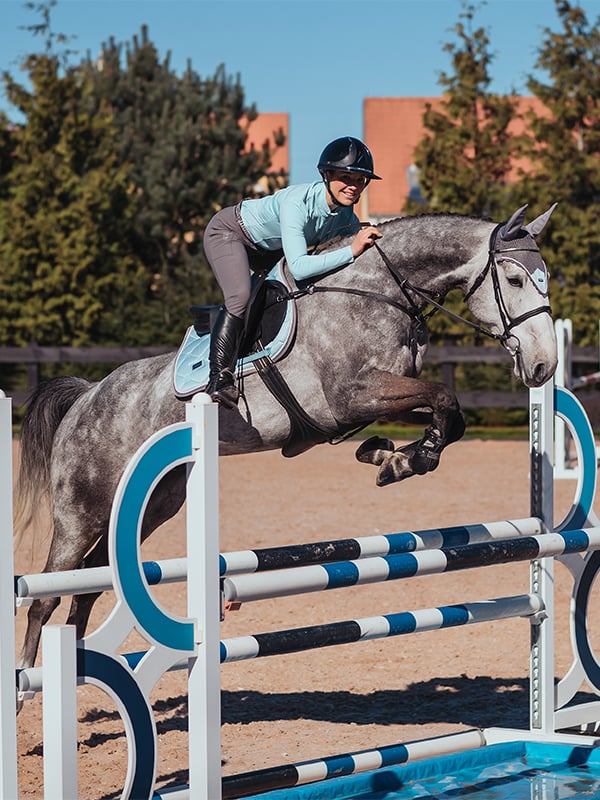 Sottosella da salto Ice Blue Equestrian Stockholm e cuffia per orecchie su cavallo grigio pomellato e cavaliere in salto.
