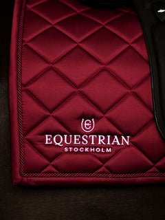Studio image of Equestrian Stockholm Dressage Saddle Pad Line Bordeaux featuring white logo embroidery and rope piping.