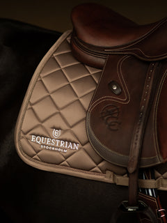 Studioaufnahme der Equestrian Stockholm Springschabracke Line Champagne mit braunem Ledersattel auf dunklem Pferd.