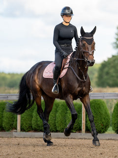 A rider in black attire on a bay horse wearing the Equestrian Stockholm Jump Saddle Pad Line Pink in an outdoor arena.