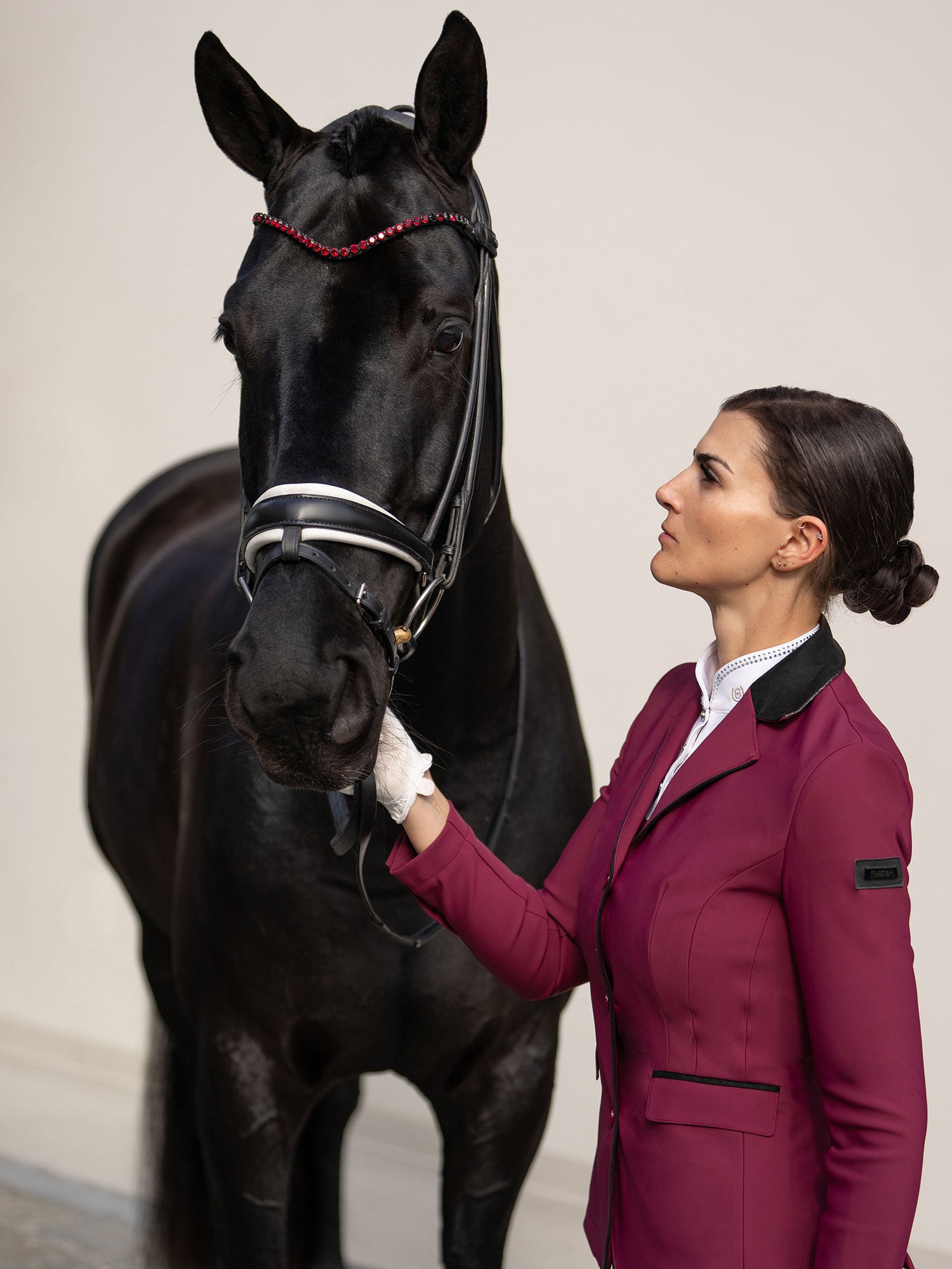 Cavalière en veste de concours Classic Merlot Equestrian Stockholm aux côtés d'un cheval noir au frontal rouge assorti.