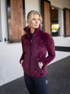 Une cavalière porte une veste polaire Velvet Merlot Equestrian Stockholm et un legging noir dans une écurie.