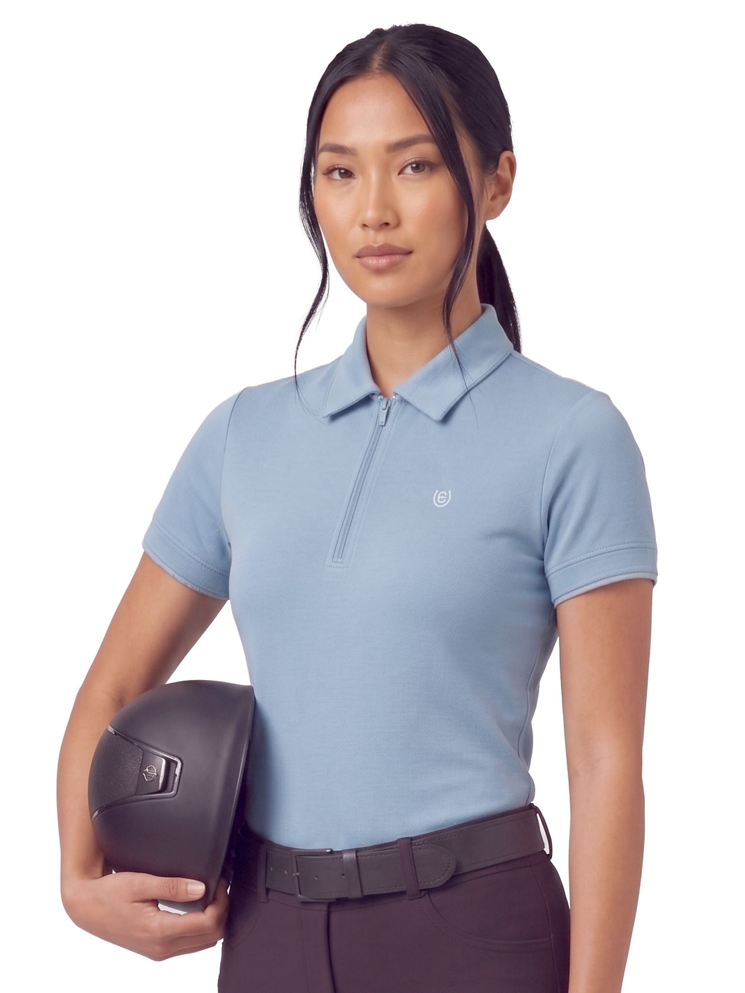 A model wearing the Equestrian Stockholm Short Sleeve Zip Top in Soft Blue holding a black riding helmet.