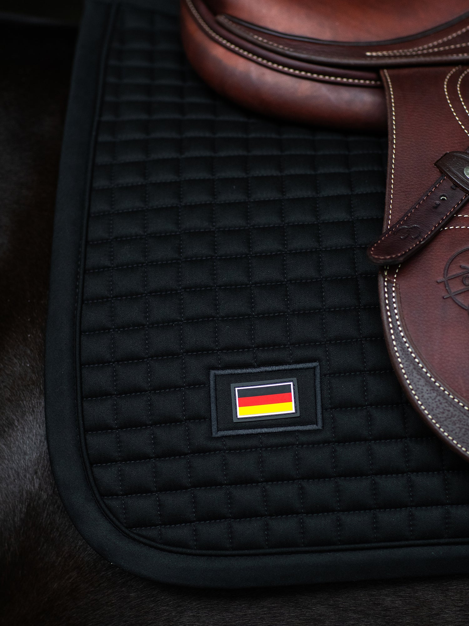 Equestrian Stockholm Black Germany Jump Saddle Pad with square quilting and a flag patch under a brown leather saddle.